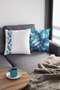 Two Sea Otter Tie-Dye pillows, front and back, on a grey couch in a bright living room