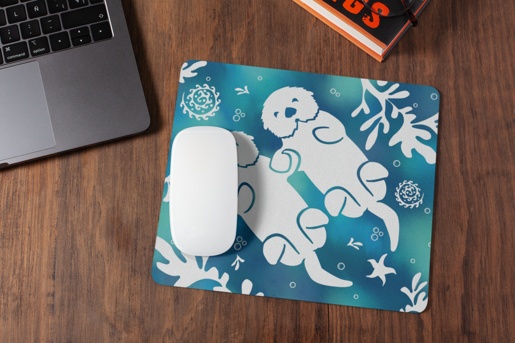 A white computer mouse sits on the Sea Otter Tie Dye Mouse Pad, which features two otters holding hands among seaweed, starfish, and shells, placed on a wooden desk next to a laptop and books.