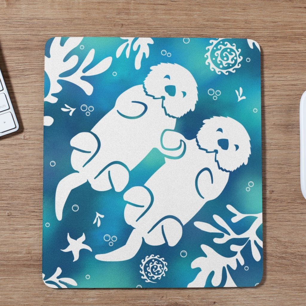 A mousepad featuring two white illustrated otters holding hands, surrounded by seaweed, starfish, and shells on a blue and green gradient background. The mousepad is on a wooden desk next to a keyboard and mouse.