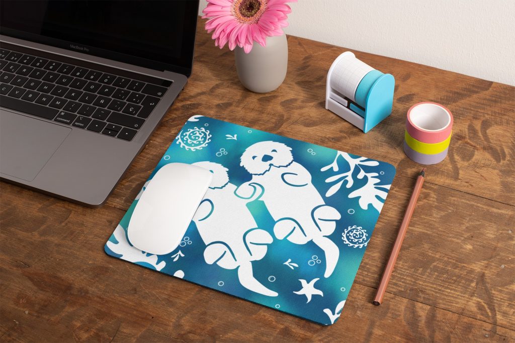A wooden desk with a laptop, a pink pencil, a roll of rainbow-striped tape, a tape dispenser, a vase with a pink flower, and a mouse on a blue mouse pad featuring two white otters surrounded by sea-themed designs.