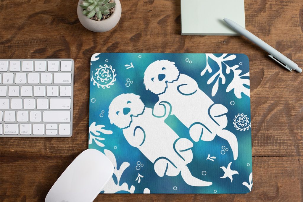 Mousepad-mockup-lying-on-a-table-next-to-a-plant-pot-and-some-office-supplies-27553 | Otter Things A mouse pad with a design of two white otters holding hands, surrounded by aquatic plants and swirls, sits on a wooden desk next to a keyboard, mouse, pen, notepad, and a small potted succulent.