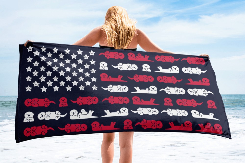 A woman standing on a beach holds a towel designed like the American flag, with stars and red-and-white stripes made up of cartoon dogs in various poses.