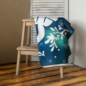 The Sea-Otter-Tie-Dye-Beach-Towel in blue and green with white snowflake and dot patterns is draped over a simple wooden stool on a rustic wooden floor, beside a white louvered panel and a closed door.