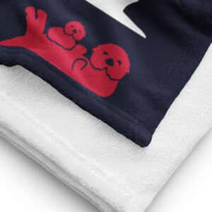 A close-up of a folded Sea Otter USA Flag Beach Towel with a navy blue and white design featuring two red sea otters, one larger and one smaller, lying down. The towel has a textured white edge.