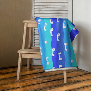 The Ocean Otter Gradient Beach Towel with blue and turquoise hues, featuring white otter designs, is draped over a simple wooden chair on a rustic wooden floor beside a louvered door.