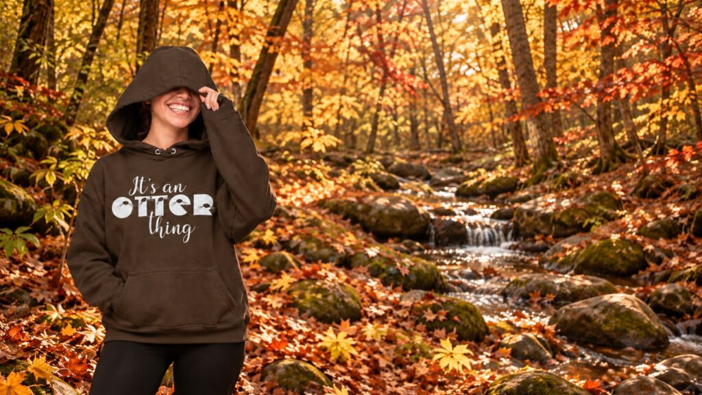 A smiling person wearing an It's An Otter Things hoodie in dark chocolate, standing in a forest with autumn leaves and a small stream.