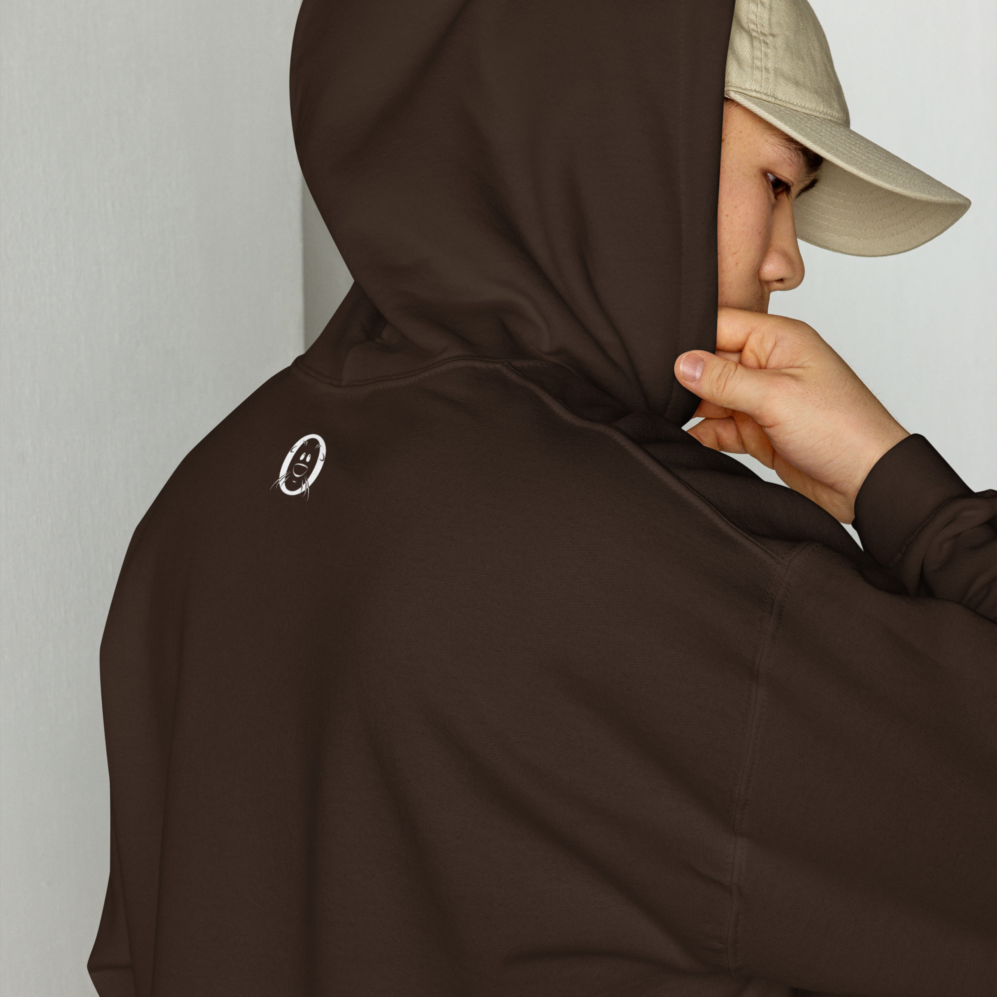 Unisex-heavy-blend-hoodie-dark-chocolate-back-6abac5d643ee8 | Otter Things Person wearing a brown hoodie with a small white smiley face on the back and a beige cap, facing sideways and pulling the hoodie’s hood closer to their face.