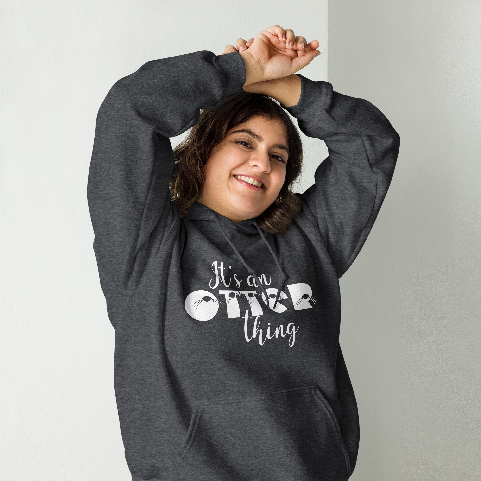 Unisex-heavy-blend-hoodie-dark-heather-front-6abac5d643751 | Otter Things A smiling person in a dark hoodie with the text “It’s an Otter thing” stretches their arms above their head, standing against a plain white wall.